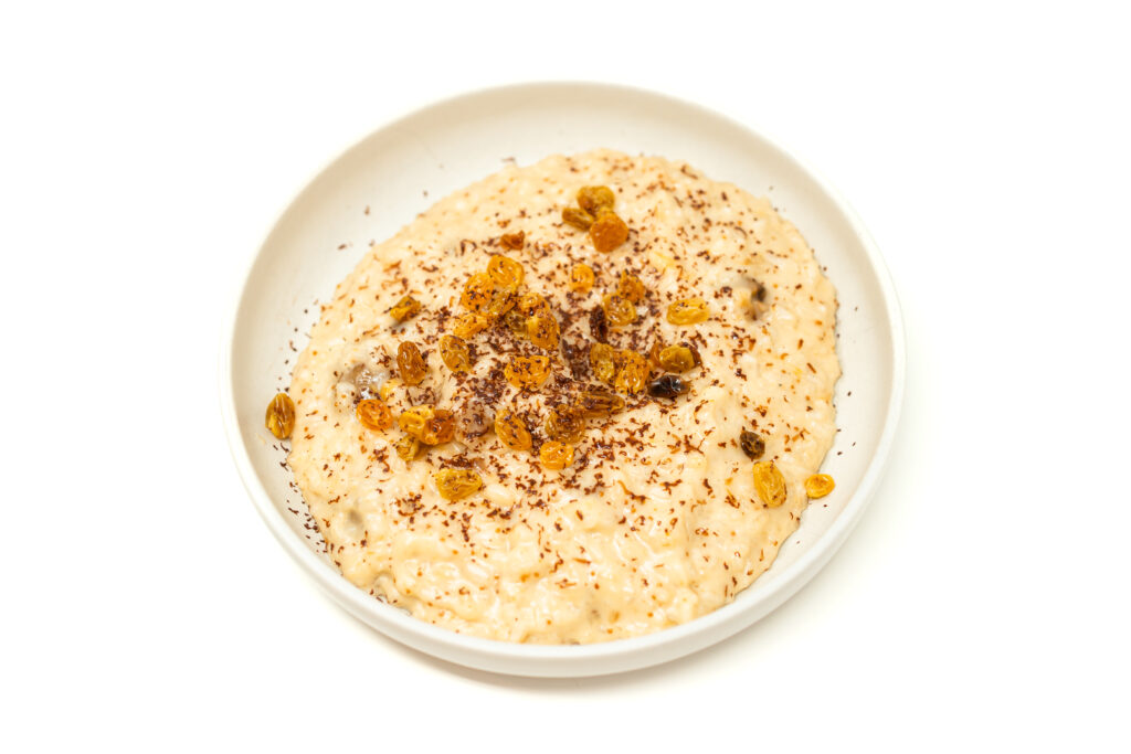 A close-up overhead shot of a white bowl filled with creamy rice pudding garnished with golden raisins and dark chocolate shavings on a white background.