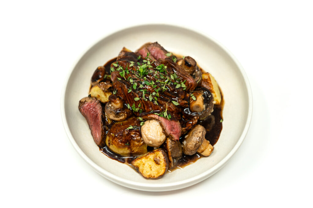 Overhead view of sliced medium-rare top sirloin steak topped with dark brown beer gravy, served in a white bowl over roasted potatoes and whole sautéed mushrooms.