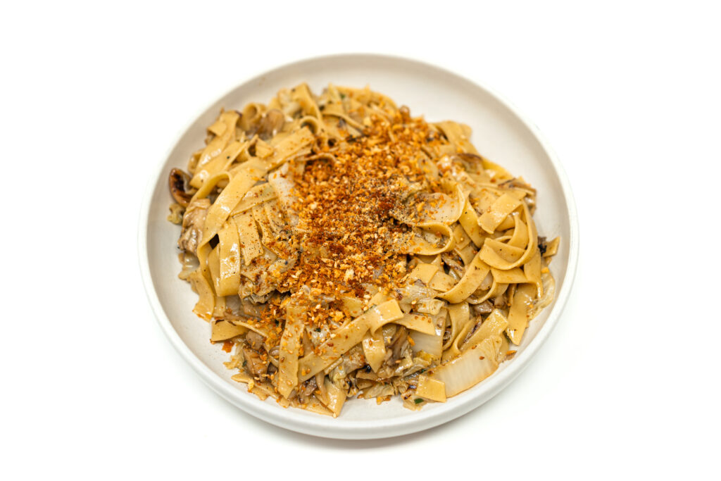 A white bowl containing wok-charred maitake mushroom and napa cabbage tagliatelle pasta tossed in a soy butter sauce and topped with golden toasted sesame panko breadcrumbs.