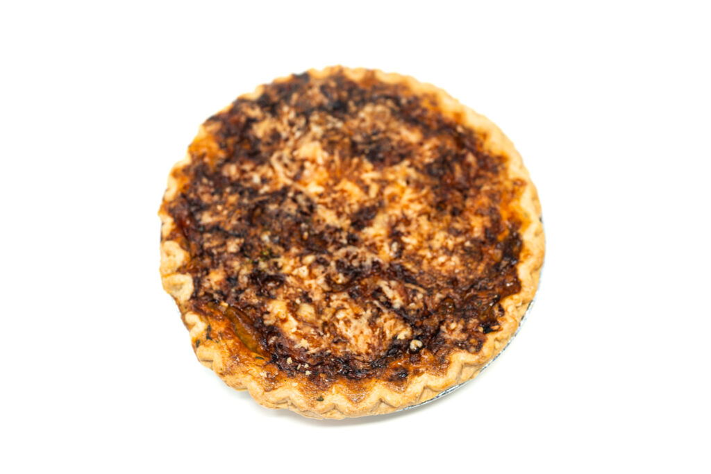 A slice of golden brown caramelized onion and Swiss cheese tart on a plate, showing layers of mustard-coated crust, dark jammy balsamic onions, and creamy set custard.
