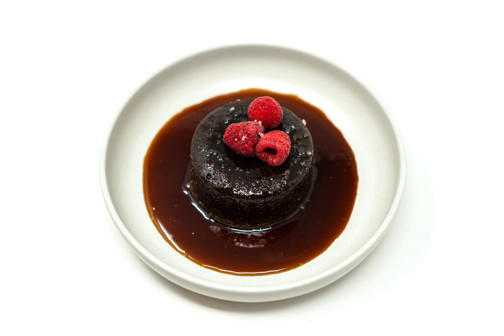 A dark, dense Valrhona chocolate custard cake on a white plate, pooled with amber Riesling caramel sauce and topped with fresh red raspberries.