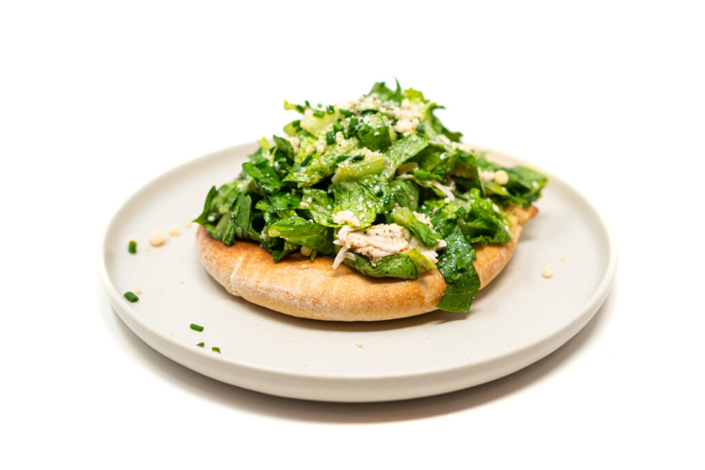 Romaine lettuce and shredded rotisserie chicken in caesar dressing piled on a toasted pita bread on a white ceramic plate.