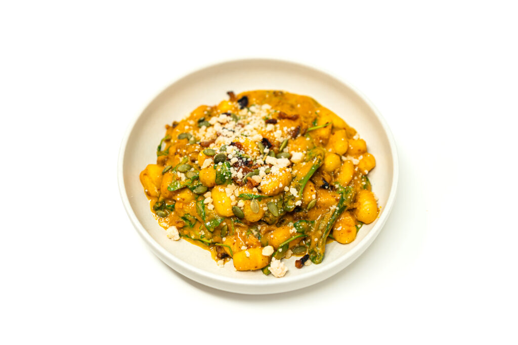 A white bowl filled with potato gnocchi coated in a vibrant orange calabaza squash cream sauce topped with blistered shishito peppers, crispy bacon, arugula, and toasted pepitas.
