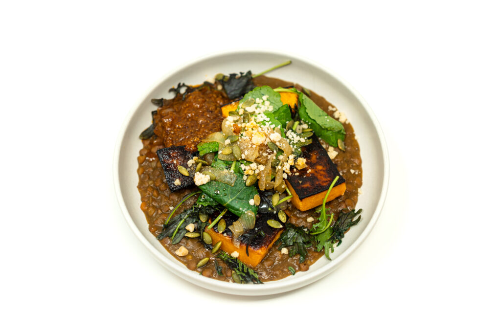 A shallow white bowl containing thick lentil soup topped with two charred butternut squash planks, fresh baby greens, a savory apple-onion relish, and grated Parmesan cheese on a white background.