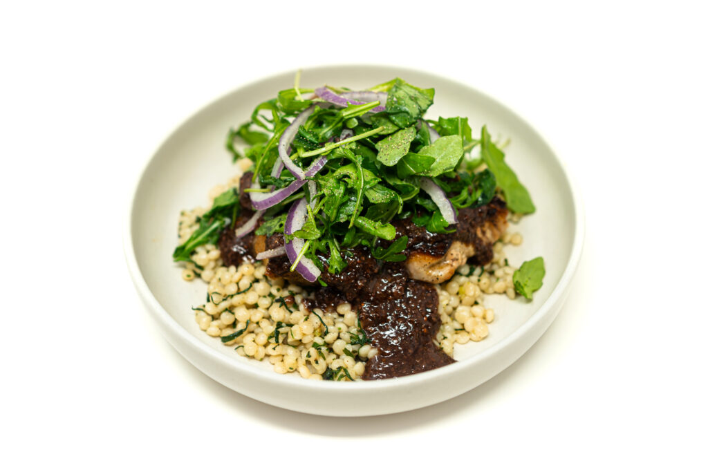A gourmet plated seared lamb chop smothered in a dark mustard wine sauce over lemon herb pearl couscous and topped with fresh arugula.
