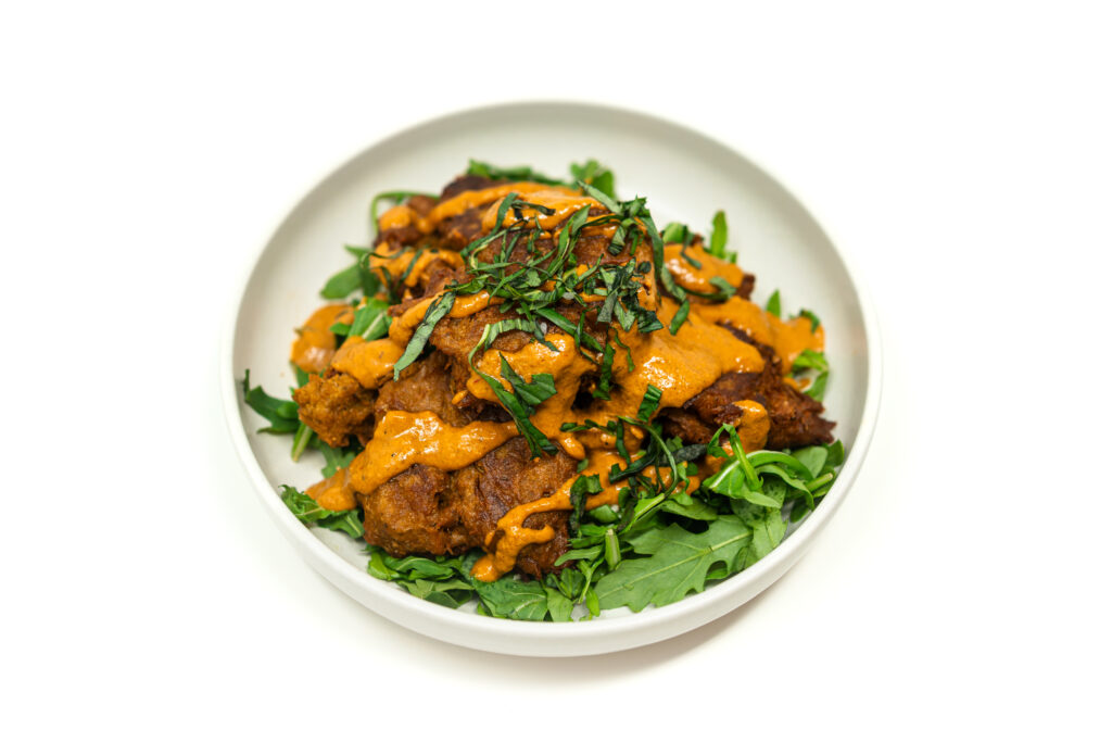 A white bowl filled with golden brown vegetable scrap fritters served over a bed of fresh green arugula and drizzled with a creamy orange sauce.