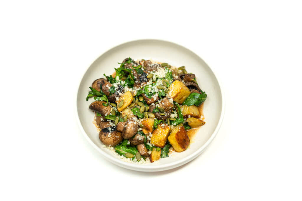 A white bowl featuring roasted Yukon gold potatoes, button mushrooms, and fresh arugula topped with Parmesan, toasted pepitas, and a dark balsamic-like broth reduction.
