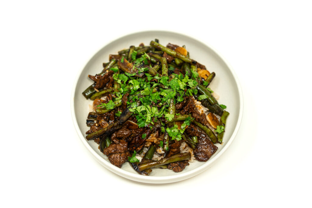 A white bowl filled with glossy black pepper beef, charred green beans, and fingerhot peppers topped with fresh cilantro.