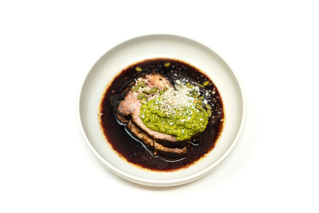 Sliced roasted pork sirloin medallions fanned over a dark red wine reduction and topped with bright green tomatillo salsa verde and crumbled Cotija cheese on a white plate.