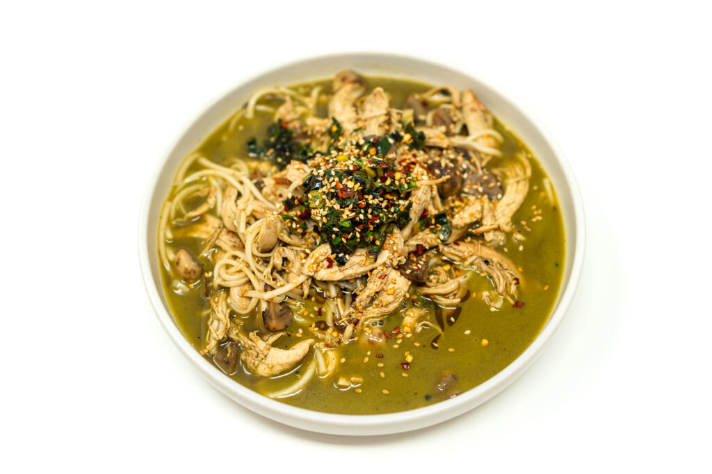 Top-down view of a bowl of creamy green miso leek soup filled with noodles, shredded chicken, and mushrooms, garnished with sesame seeds and chili oil.