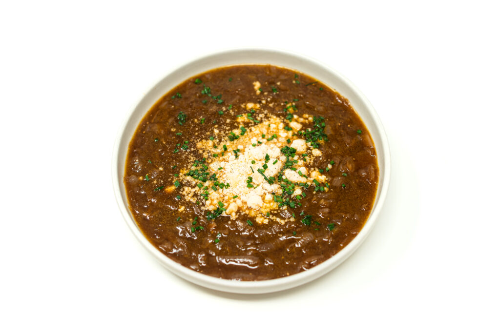 Top down view of a dark mahogany red wine onion soup in a white bowl, topped with a mound of fluffy grated Parmesan cheese and fresh chopped chives.