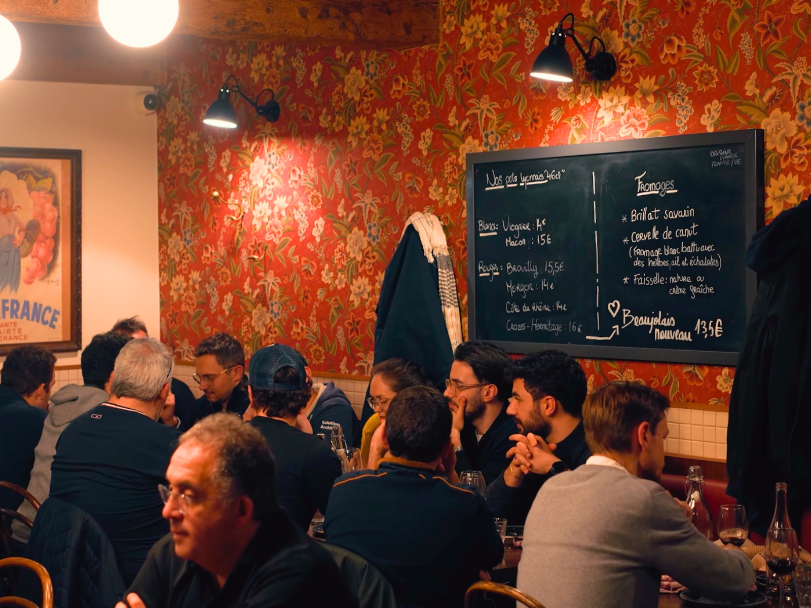 The bustling dining room of Le Bouchon des Filles featuring red patterned wallpaper and a chalkboard menu.