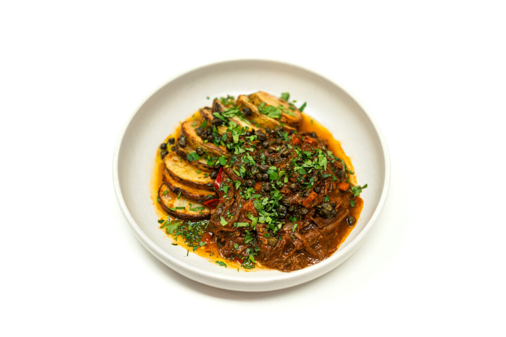 A white bowl filled with rich brisket ropa vieja topped with crispy fried capers and fresh cilantro alongside golden roasted potato rounds.