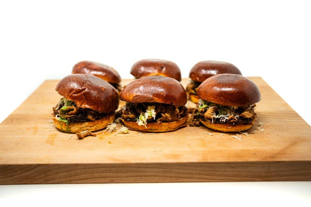 Six shaved chili pork sliders on a wooden board, featuring melted white cheddar, dark onion jam, and bright green herb pesto on toasted brioche buns.