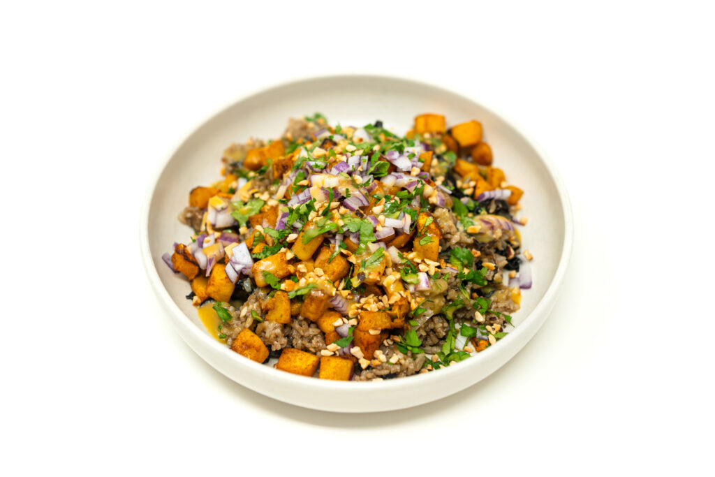 A ceramic bowl filled with dark wild mushroom jasmine rice, roasted butternut squash cubes, diced red onion, and crushed hazelnuts.
