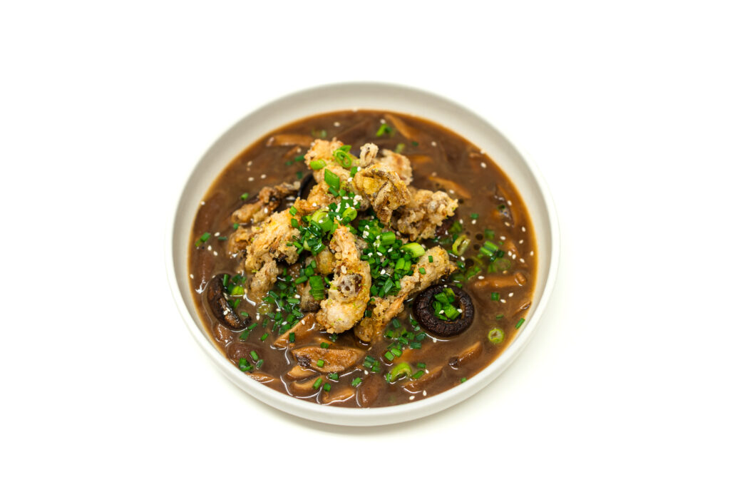 Top-down view of dark brown mushroom soup in a ceramic bowl topped with a pile of golden fried oyster mushroom tempura and scallions.