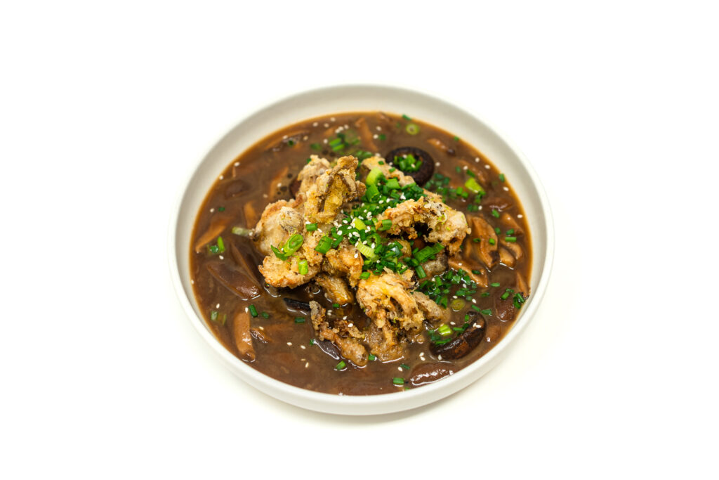 Top-down view of dark brown mushroom soup in a ceramic bowl topped with a pile of golden fried oyster mushroom tempura and scallions.