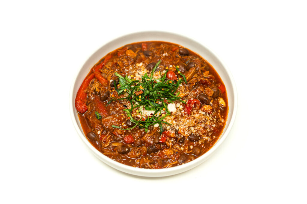 Overhead shot of tomato-braised pork bone chili in a white bowl, garnished with fresh chiffonade herbs, grated Cotija cheese, and red pepper flakes.