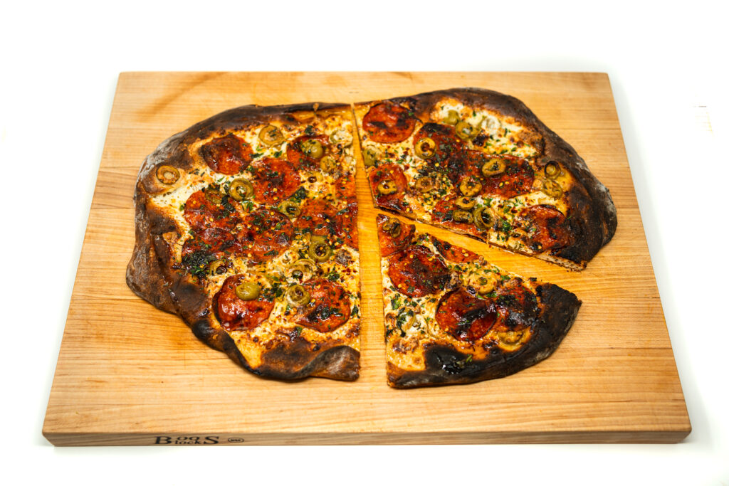 A rustic white pizza on a wooden cutting board topped with melted fresh mozzarella balls, spicy pepperoni, and green olives with charred crust edges.