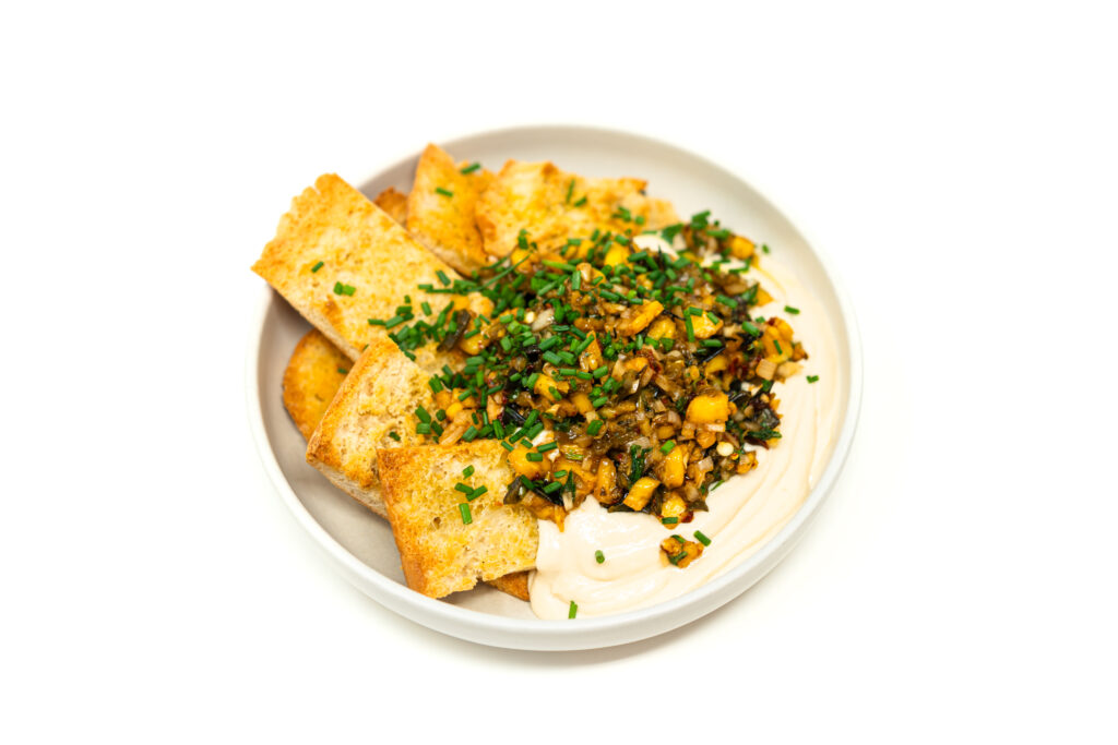 A white bowl containing diced pineapple and roasted poblano salsa on a bed of whipped garlic tahini spread with toasted naan bread.