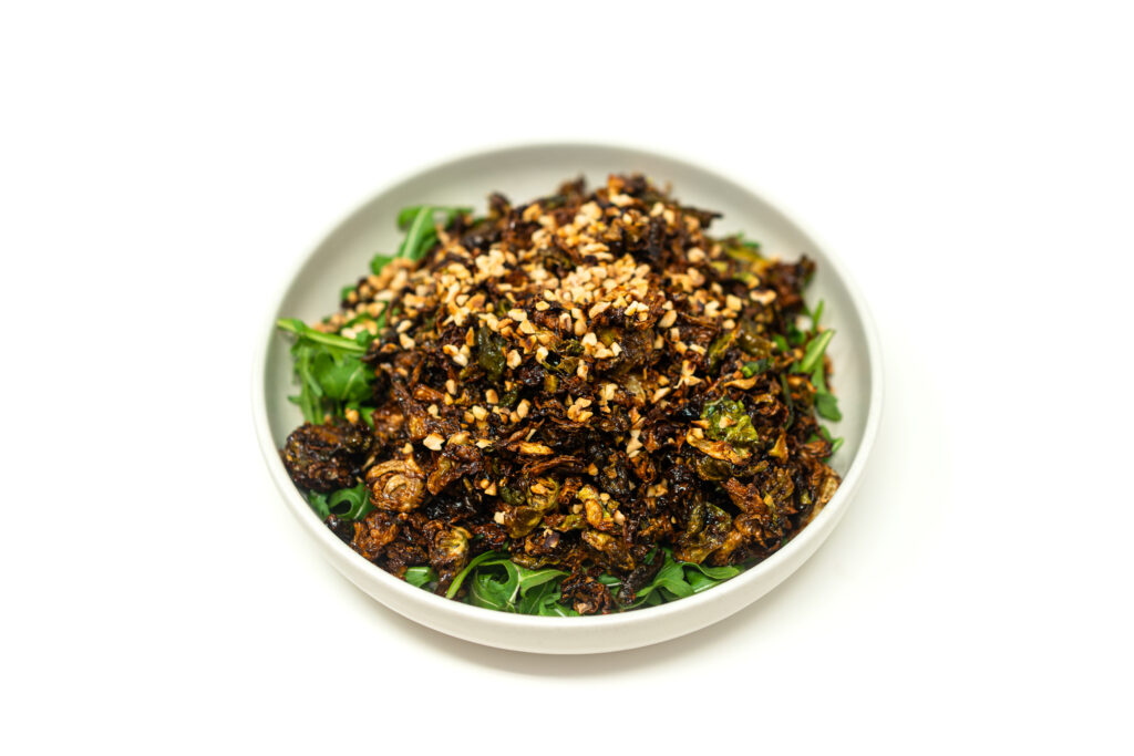 A white bowl filled with crispy fried sliced brussels sprouts and shishito peppers coated in a dark berry glaze and topped with crushed hazelnuts.