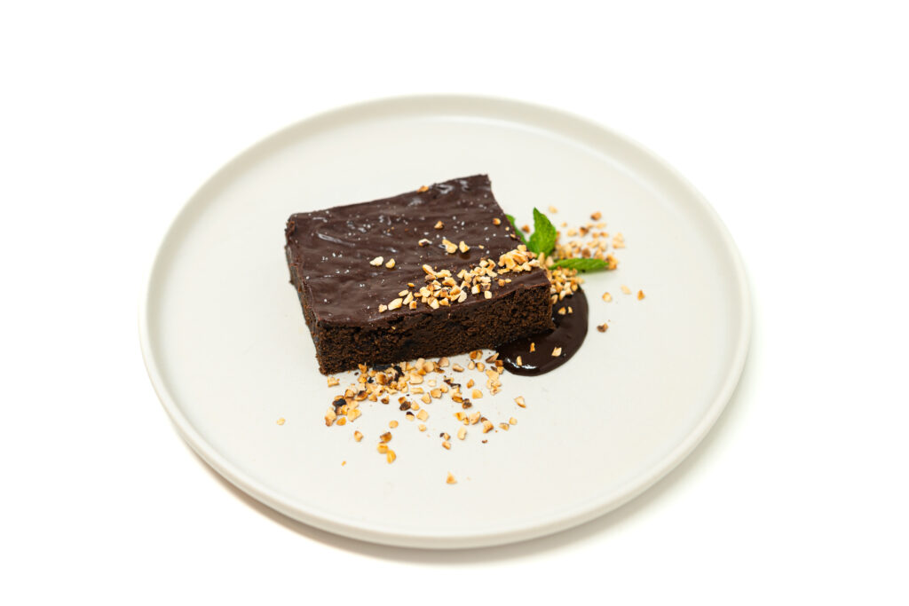 A thick slice of chocolate blackberry sheet cake on a white plate topped with dark ganache, crushed hazelnuts, and fresh mint.