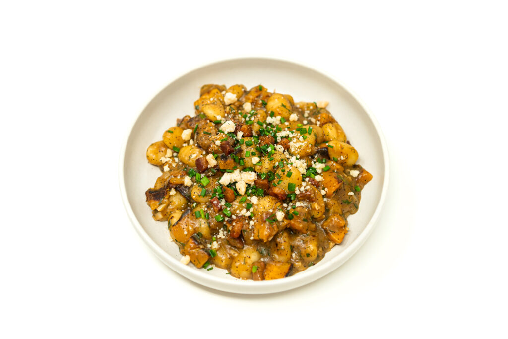 A shallow bowl filled with seared gnocchi with acorn squash, crispy pancetta bits, and chopped chives over a rich, glossy miso butter sauce.