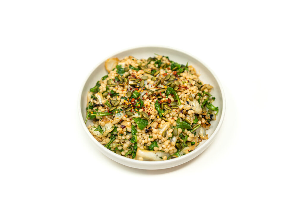 A white bowl filled with charred cabbage pearl couscous salad topped with arugula, toasted pepitas, and red pepper flakes.