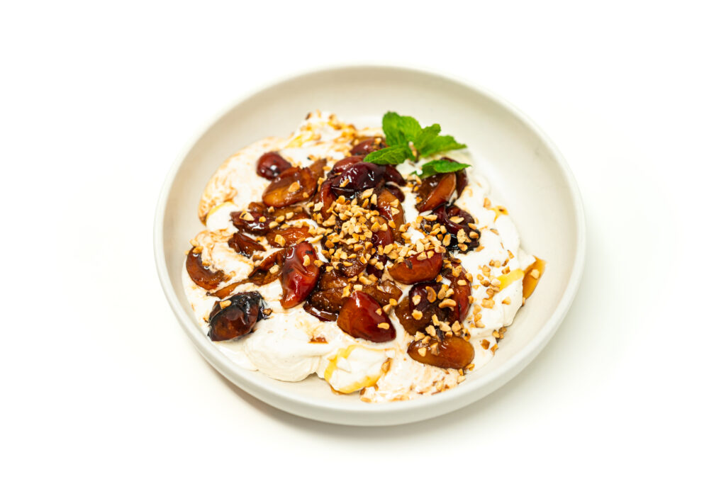 A wide bowl filled with soft vanilla whipped cream topped with warm roasted balsamic plums, toasted hazelnuts, honey, and fresh mint.