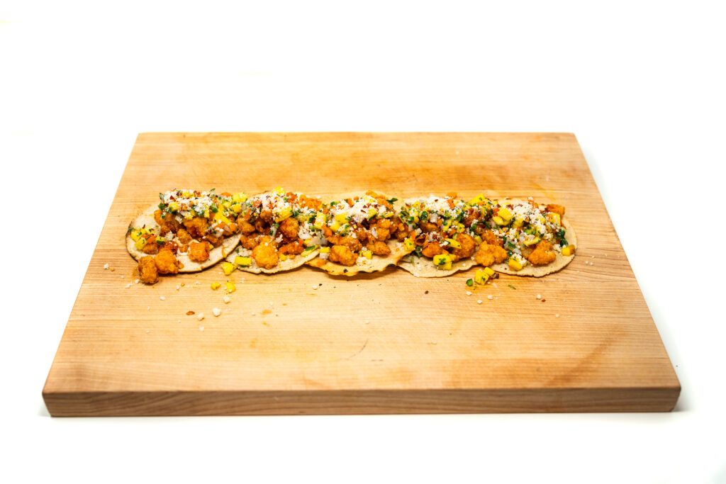 Wooden board lined with popcorn shrimp tacos topped with mango caper salsa and cotija cheese.