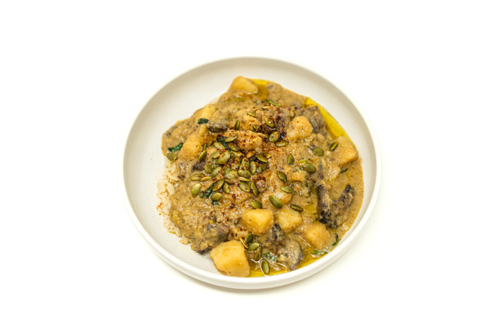 A shallow bowl of rich braised beef neckbone and potato green curry topped with toasted pepitas, chili oil, and served over white rice.