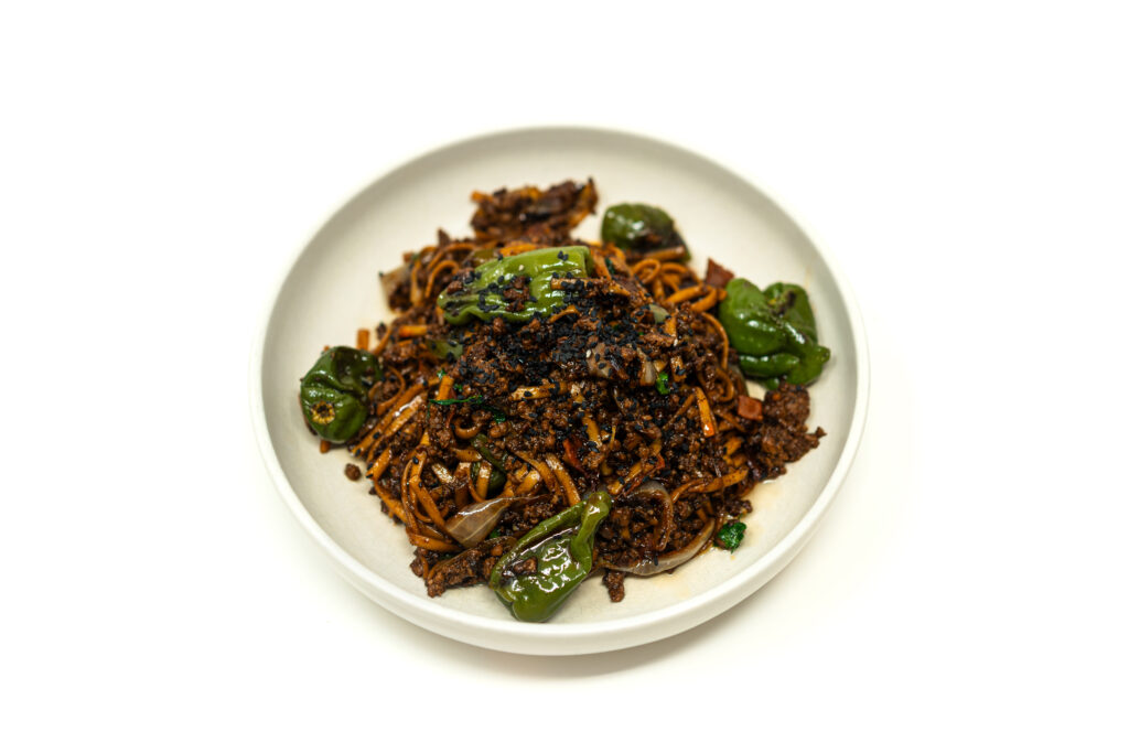 Overhead view of dark glossy udon noodles mixed with ground beef, chopped pepperoni, and blistered green shishito peppers in a white bowl.