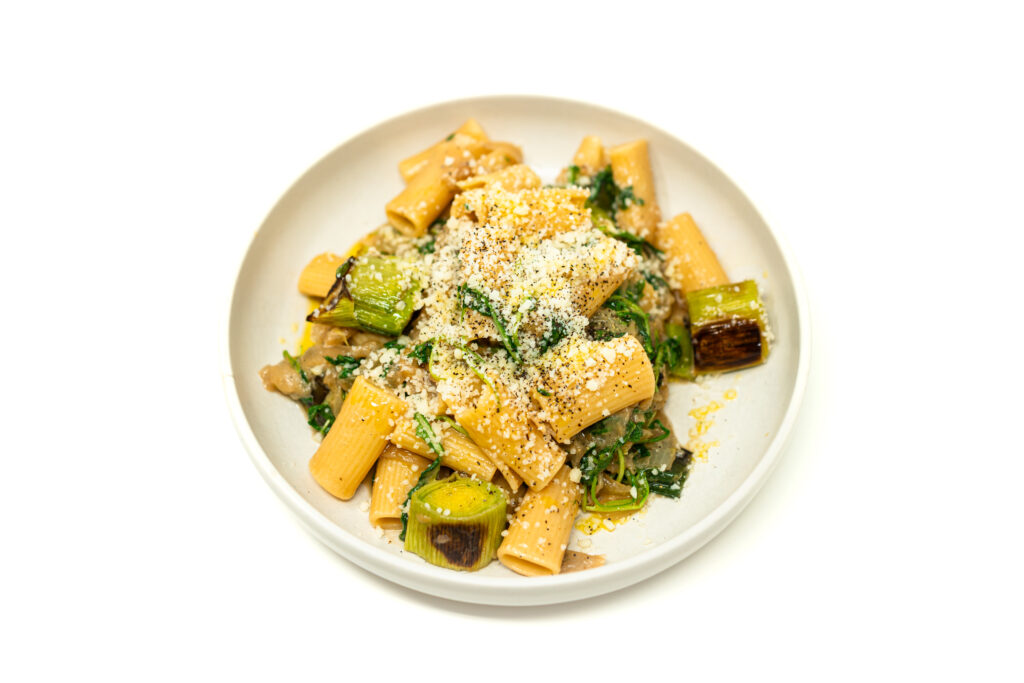 A wide bowl of rigatoni coated in caramelized onions and asiago cheese, topped with charred leek medallions and wilted arugula.