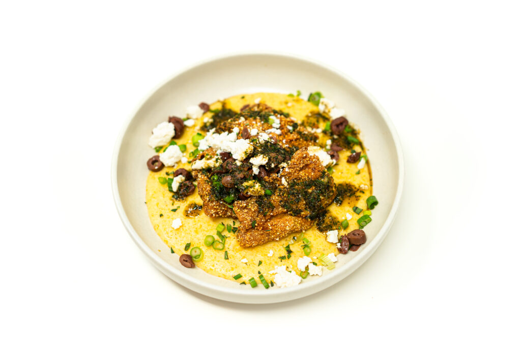 A shallow bowl filled with creamy yellow polenta, topped with golden crispy fried fish pieces, green sauce, crumbled white cheese, and chopped olives.