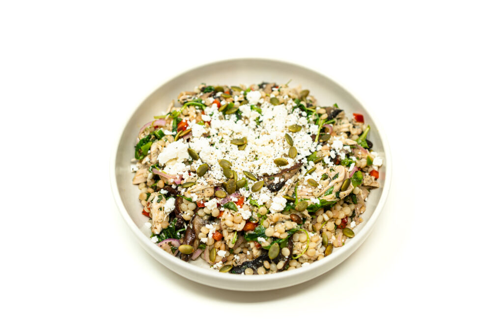 A top-down view of a white bowl filled with shredded chicken and pearl couscous salad, topped with crumbled cheese and pumpkin seeds.