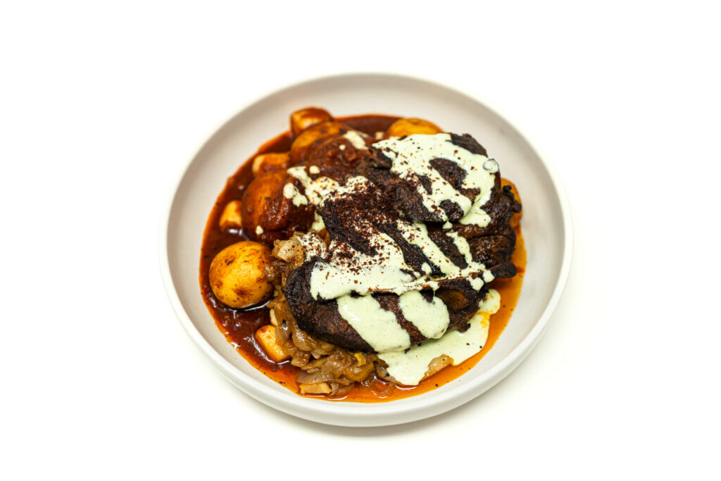 Plate of crusty braised lamb shoulder steak resting on red tomato potatoes with a drizzle of green herb crema and sumac.