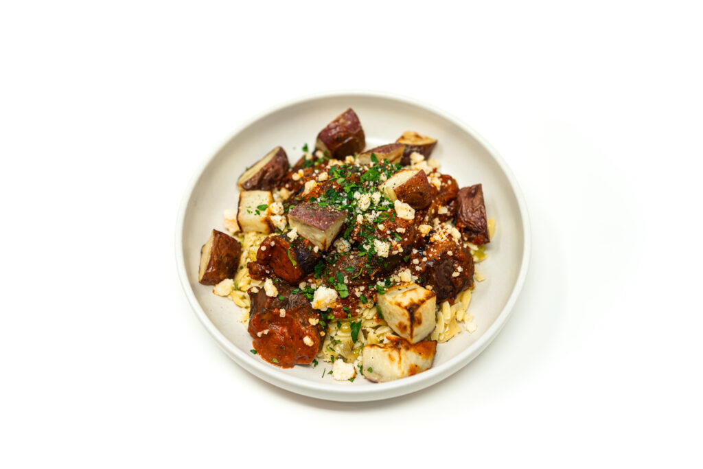 A shallow white bowl containing tomato-braised Chinese eggplant served over creamy leek orzo and topped with crispy roasted red potatoes.