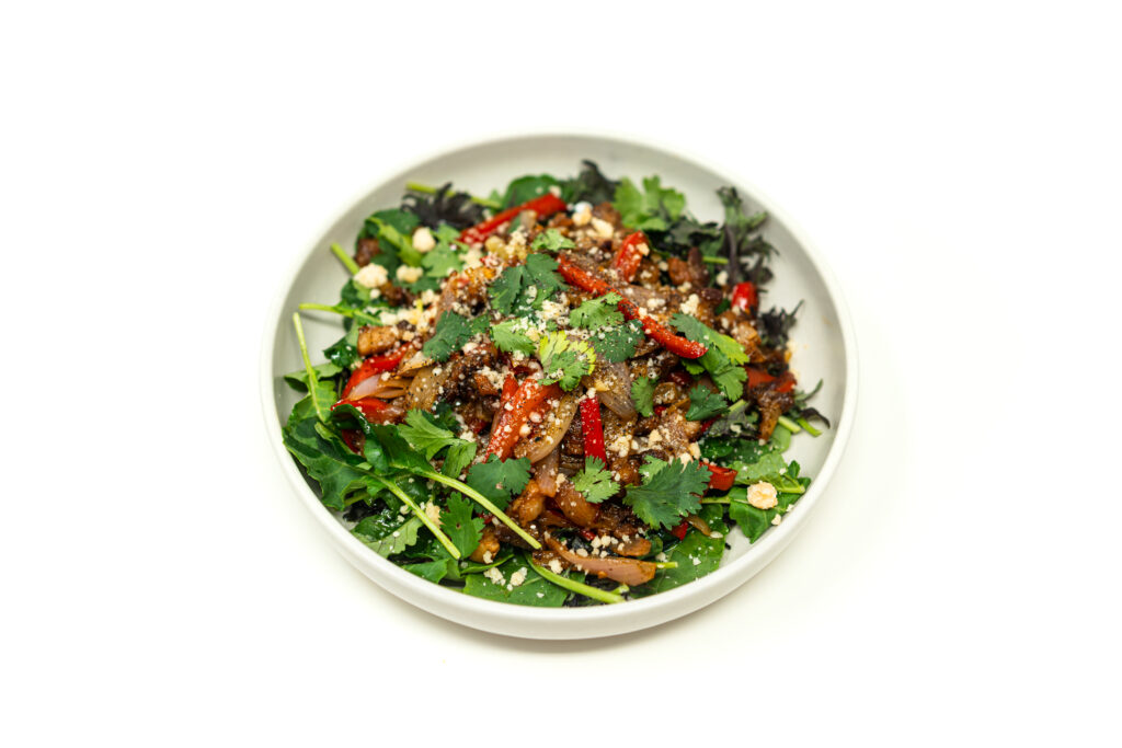 A white bowl filled with hard seared mezcal glazed chicken, peppers, and onions over a bed of dark greens, topped with cotija and cilantro.