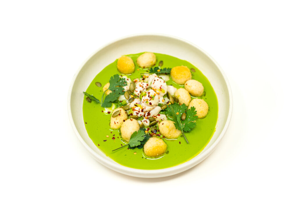 A wide shallow bowl holding bright green aguachile sauce, topped with diced scallop ceviche, seared masa gnocchi, and toasted pepitas.