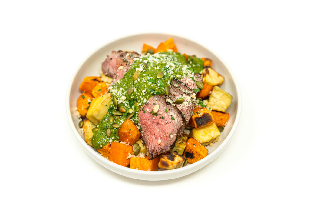 A sliced soy-brined tri-tip steak resting on roasted root vegetables, topped with vibrant green blended herb chimichurri, cotija cheese, and toasted pepitas.