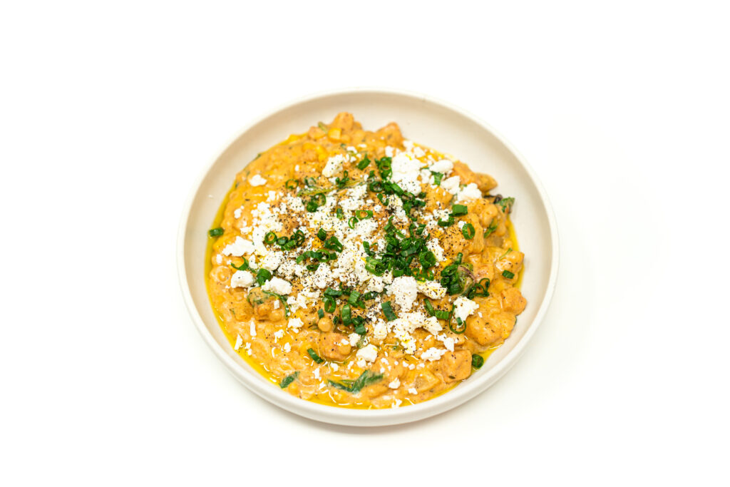 A wide bowl of golden pearl couscous congee topped with crumbled feta, fresh green onions, and a drizzle of sizzling ginger jalapeño avocado oil.