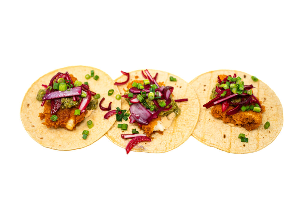 Three charred corn tortillas filled with crispy golden fried fish, green salsa, and bright magenta pickled onions on a stark white surface.