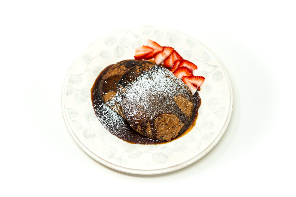 A stack of dark brown plantain pancakes drenched in thick espresso syrup, dusted heavily with powdered sugar, and served with sliced strawberries on a white plate.