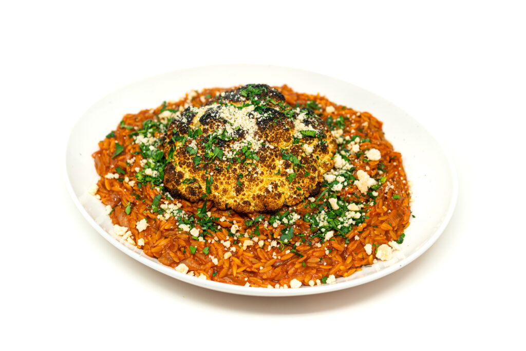 A heavily charred, spice-rubbed head of cauliflower resting over a rich red tomato and caramelized onion orzo pasta, garnished with parsley.