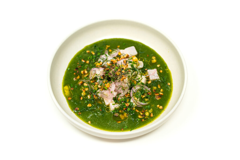 A shallow bowl filled with vibrant green basil-dill aguachile, centered with cubed corvina ceviche, topped with charred corn and olive oil.