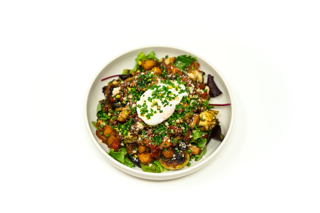 Overhead view of a bowl containing miso-honey brussels sprout hash topped with a poached egg and chives over mixed greens.