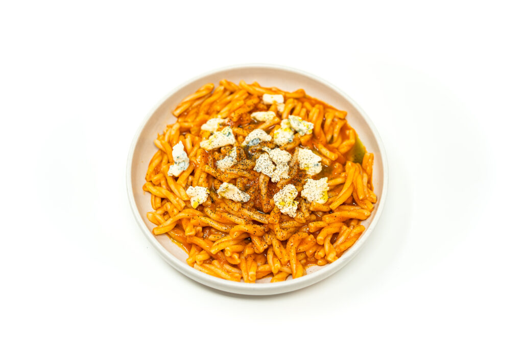 A white bowl filled with short pasta coated in a vibrant chipotle rosé tomato sauce, topped with crumbled gorgonzola and bright green herb oil.