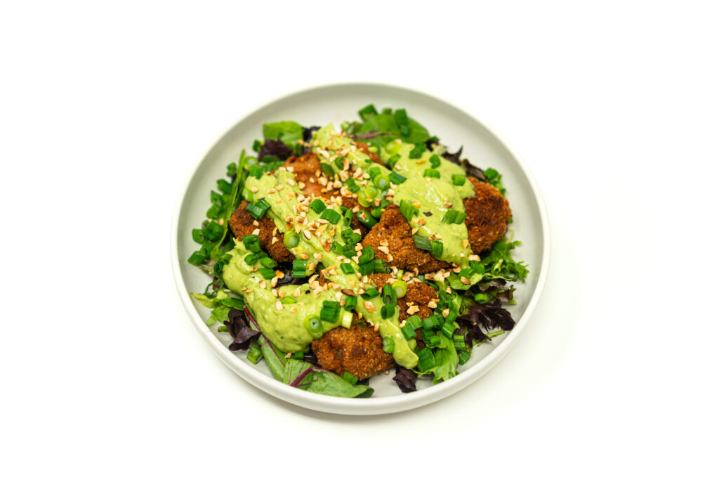 Golden brown chunks of pan fried chicken heavily coated in crushed pistachios, resting on spring greens and heavily draped in a pale green avocado cilantro crema.