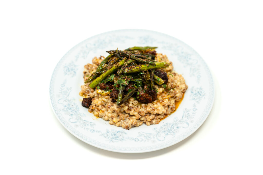 A layered plate of creamy risotto-style pearl couscous topped with charred asparagus, long beans, crispy chorizo, and kiwi vinaigrette.