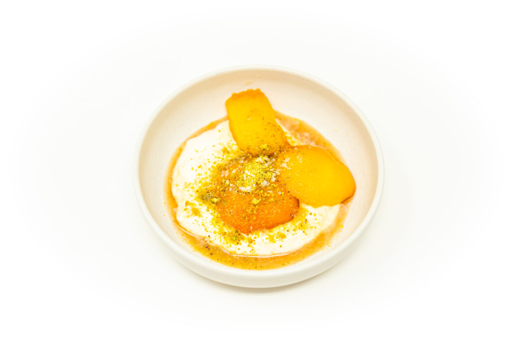 A shallow bowl filled with burnt honey yogurt cream, topped with warm poached peaches, white port syrup, and crushed pistachios.